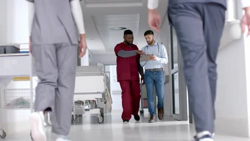 Diverse male doctors discussing work using tablet walking in hospital corridor, slow motion