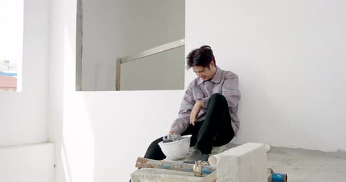 Asian male construction worker sits on unfinished concrete ledge holding helmet, pausing during