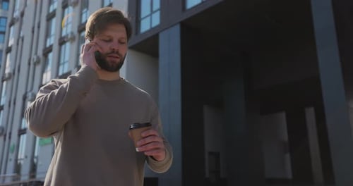 Young Bearded Man Talking on the Smartphone and Drinking Coffee at Street