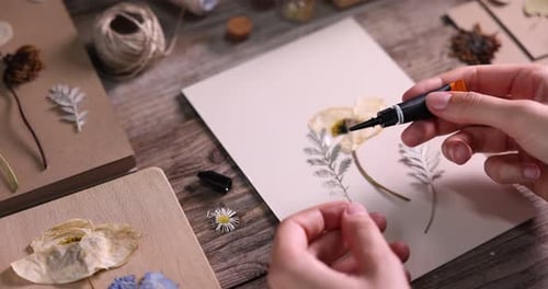 Woman putting dry flowers onto paper sheet at wooden table, closeup