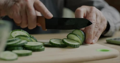 Closeup of Hands of Old Man Preparing Vegetable Salad Cutting Cucumber with Sharp Knife in Kitchen