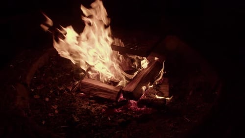 Large orange flame campfire burns logs under iron cooking grill, night