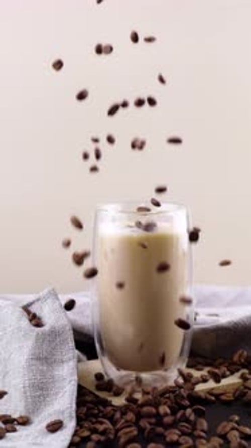Coffee beans fall into a creamy glass of coffee in slow motion, with scattered beans on a wooden boa