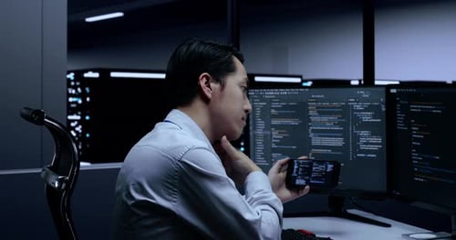 Close Up Of Male Programmer Looking At Smartphone While Writing Code By Desktops In The Server Room