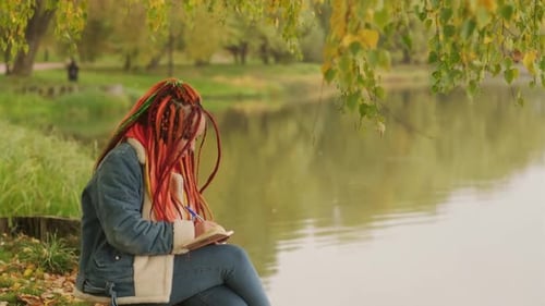 Young Creative Woman with Dreadlocks Drawing Writing in Notebook Sitting on Stumps Near Lake in
