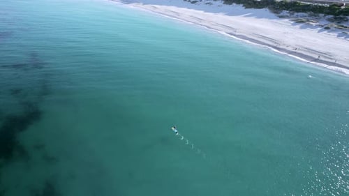 An aerial shot of a stand up paddle boarder (SUP) paddling down the coast. People can be seen on the