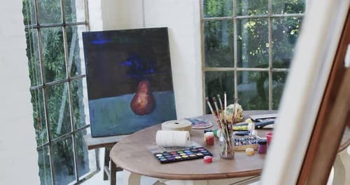 Close up of paintings on easels and paints and brushes on table in studio, slow motion