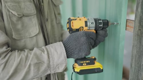 Close-up of a carpenter's hand wearing a gray glove using a drill to drill into a green metal