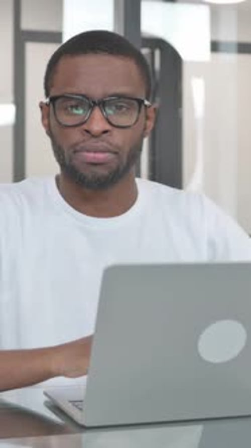 Young African Man Looking at Camera while Working on Laptop in Office, vertical video