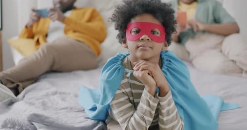 Portrait of Funny African American Boy Wearing Superhero Mask and Cape Lying in Bed with Parents