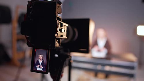 Professional camera recording a female reporter sitting at desk at blurred backdrop. Close up