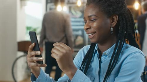 Beautiful Woman Sits in the Coffee Shop Uses Smartphone. She Smiles while Browsing Her Mobile Phone