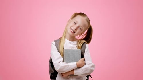 Junges Mädchen mit blonden Haaren lächelt, während es ein Buch vor rosafarbenem Hintergrund hält