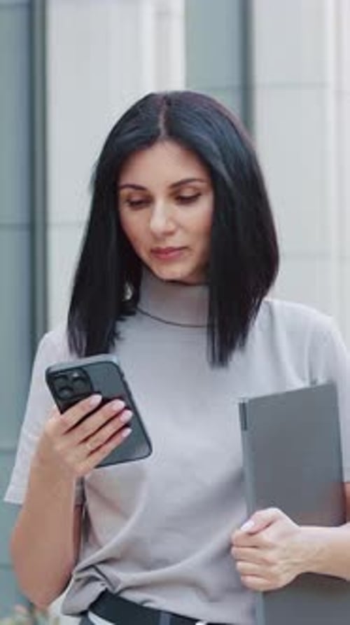 Close Up of Attractive Young with Dark Hair Woman Standing Holding Laptop and Using Phone Outdoors