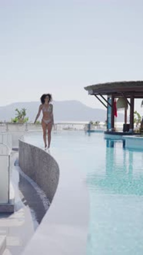 Woman in bikini walking through shallow water at the edge of an infinity pool with ocean view