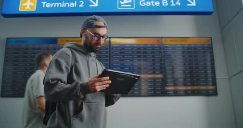 Airport Terminal Traveler Uses Tablet Computer Checks Flight Information on Digital Departure Board