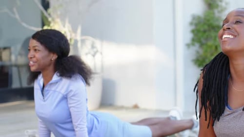 Amigas africanas en ropa deportiva sonriendo, practicando cobra en cubierta sobre colchonetas verdes con botella