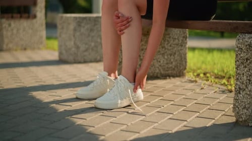 Lady Removing Sneaker Seated on Bench Outdoors with Greenery in Background