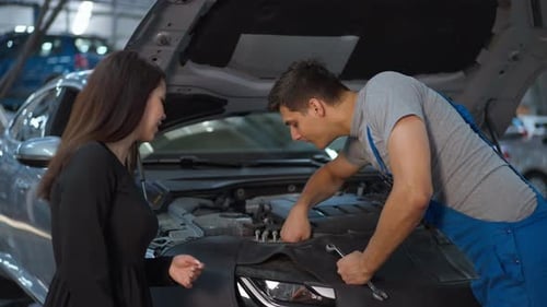 Caucasian Woman Talking with Man Examining Car Engine in Auto Service Station Indoors