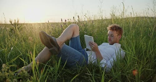 Young Male Poet Lying on Green Grass and Writing in Notepad While Creating Poem in Meadow at Sunset
