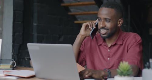 African American Employee Speaking on Mobile Phone Discussing Business and Using Laptop in Loft