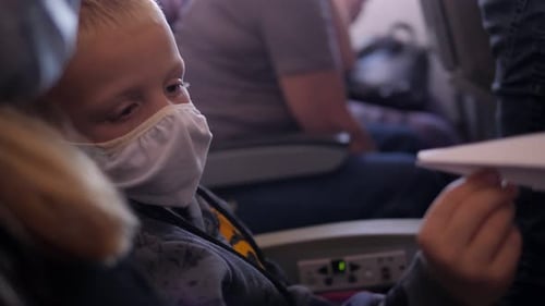 A Boy in a Mask is Playing with a Paper Airplane on an Airplane During a Flight