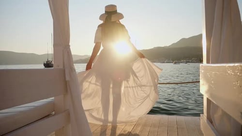 Slow motion of young brunette girl in a white dress, hat, and flowing hair and dancing on pier