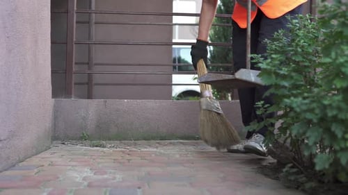A cleaning lady in a reflective vest sweeps up garbage on the street. The work of a cleaning company