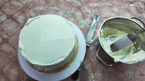 The final layer of frosting is applied to the cake. Background includes a bowl of frosting and a kni