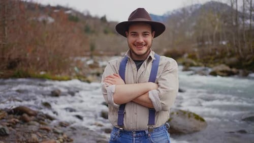 Retrato de un hombre con sombrero de vaquero y tirantes sobre el fondo de montañas y un río