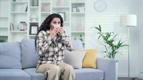 Young sad brunette woman with an allergy sneezes while sitting on sofa in living room at home. Sick