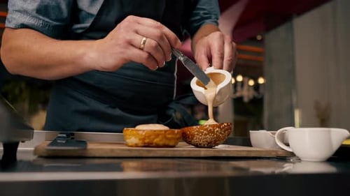 A close-up of a chef spreading sauce on a bun while cooking a burger in a professional kitchen