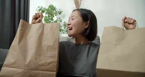 Aufgeregte asiatische Dame, die online einkauft und zu Hause Geschenktüten über ihren Kopf hebt