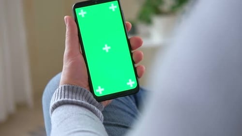 Young Man Sitting at Home Holding Smartphone Green Mockup Screen in Hand Male Person Using Chroma