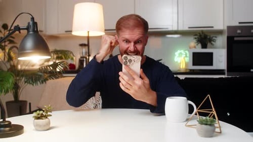 Excited man looks at phone, celebrates, sits at table