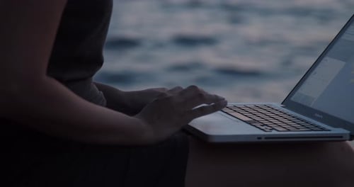 Mädchenhände mit Laptop sitzen bei Sonnenuntergang auf einem Felsen am Meer, Zeitlupe, Nahaufnahme