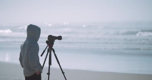 A cameraman filming or photographing the ocean from the beach in a hoodie.