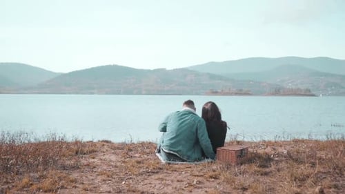 Loving Couple Sitting Near Water With Picnic Basket