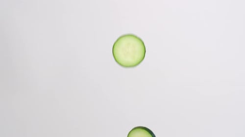 Fresh, bright green cucumber slices raining down on white backdrop in slow motion