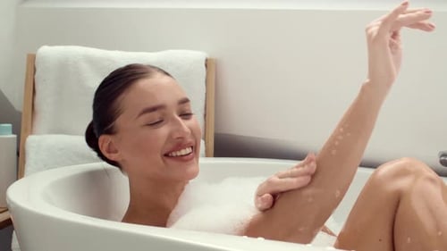 Junge unbeschwerte Dame genießt zu Hause ein entspannendes Schaumbad, spielt mit Schaum, wäscht sich die Hand und