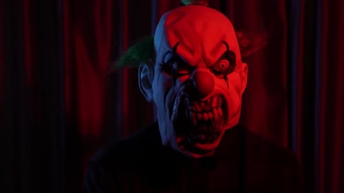 Dangerous Clown with Horror Mask in Font of Curtain Red Light