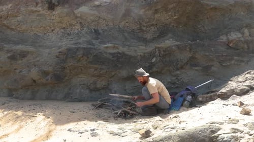 A wide shot of a bushman cooking a fish on hot coals on the coastline in Australia.