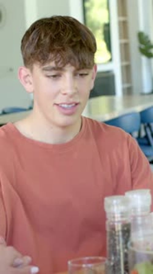 Vertical video: Listening teen in rust tee reacting while offscreen hand reaching grinders at table