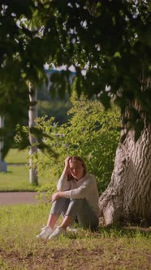 Thoughtful Woman Sitting Under Tree with Sunlight and Stadium in Background