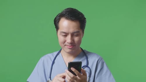 Close Up Of Asian Male Doctor With Stethoscope Using Smartphone While Standing On Green Screen