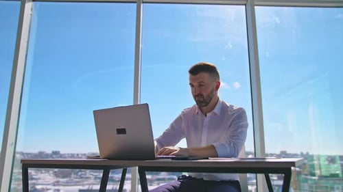 Focused businessman working on laptop computer in office with large windows.