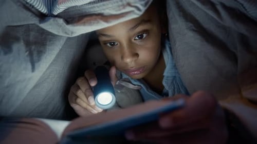 Black Girl Reading Captivating Book under Blanket with Flashlight at Night