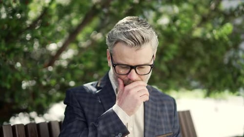 Close up of mature businessman suffers from allergies sneezes with a runny nose sitting on bench