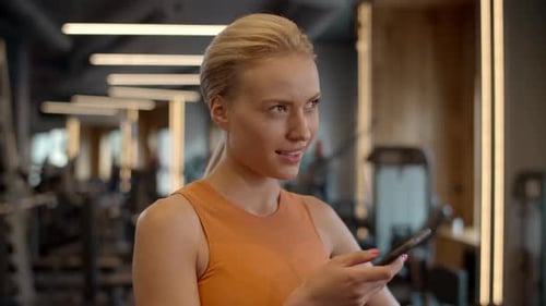 Dreamy blonde sportswoman texting on smartphone during fitness break at gym