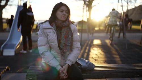 Contemplative woman seated at park bench during winter golden hour time at park, blurred children
